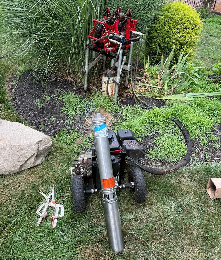 A new submersible pump on a lawn beside a red pump-pulling reel, next to a wellhead in a landscaped bed