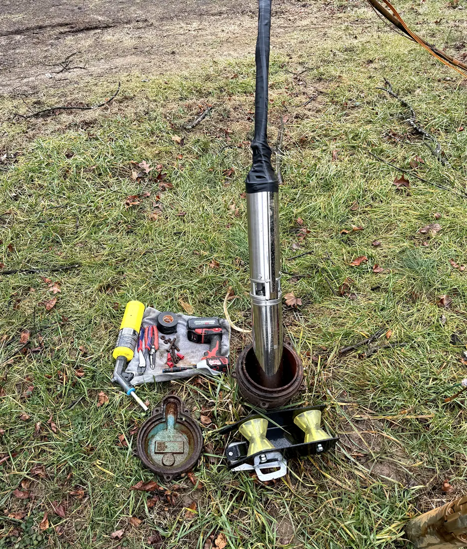 A pulled submersible pump standing in the grass beside a wellhead with tools and fittings