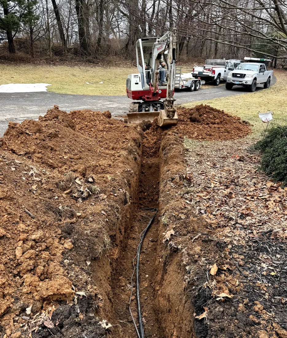 A trench across a lawn with a mini excavator and work trucks behind it